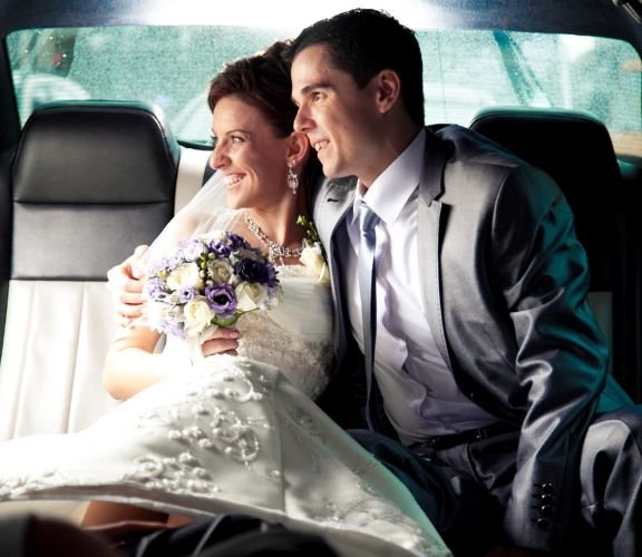 A newly married couple smiling and looking out the window from the back seat of a limousine.