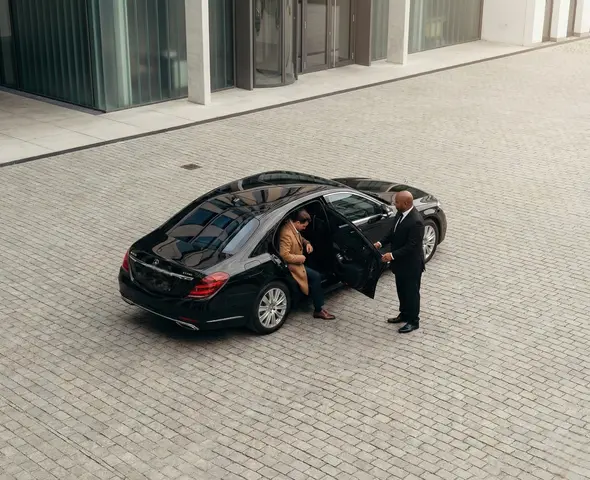 Chauffeur in a dark suit holding open the rear door of a black luxury sedan for a businessman in a tan jacket who is stepping into the car in a paved urban area.