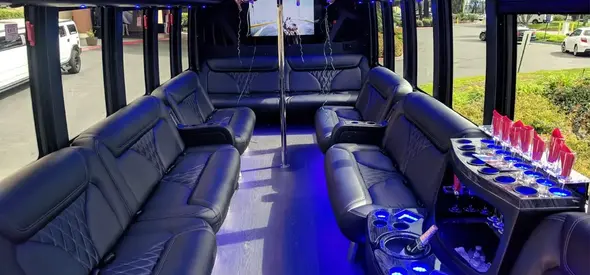 Interior of a luxury party bus featuring perimeter leather seating with diamond stitching, blue LED accent lighting, a dance pole, and a built-in bar with glassware.