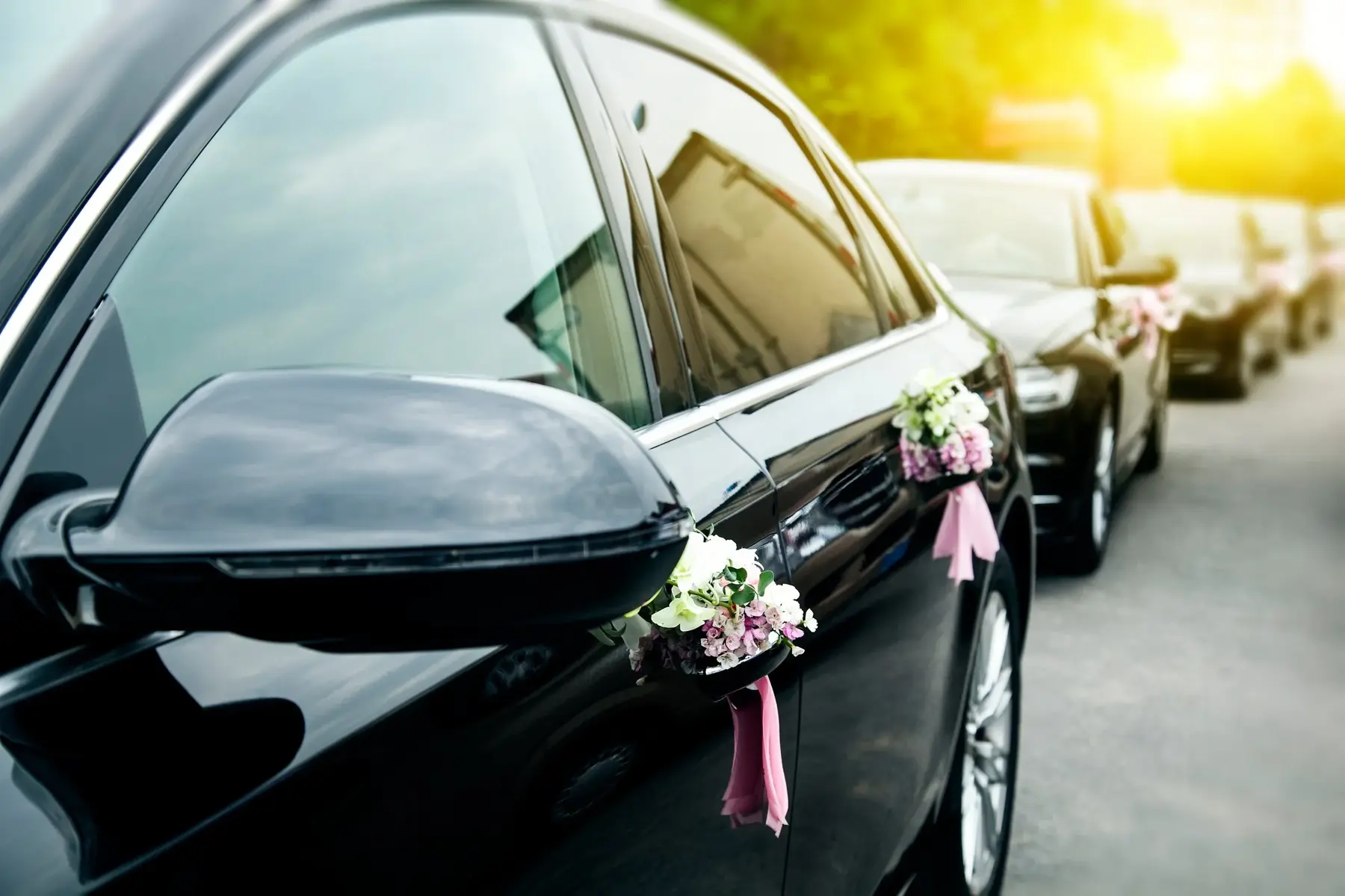 A black wedding car decorated with floral bouquets and pink ribbons on the door handles.