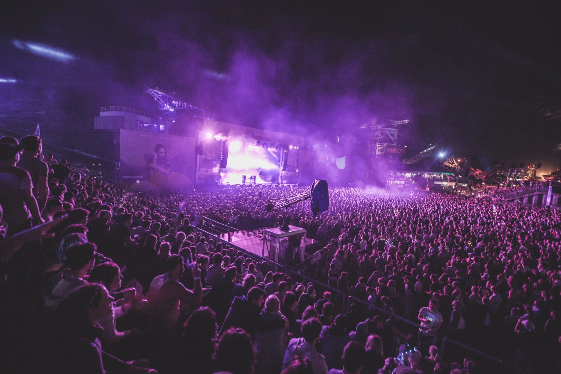 Large crowd at outdoor concert with stage lights and purple smoke at night.